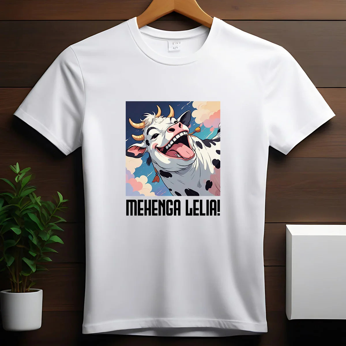 White t-shirt with colorful cartoon happy cow and text 'MEHENGA LELIA!' on wooden hanger